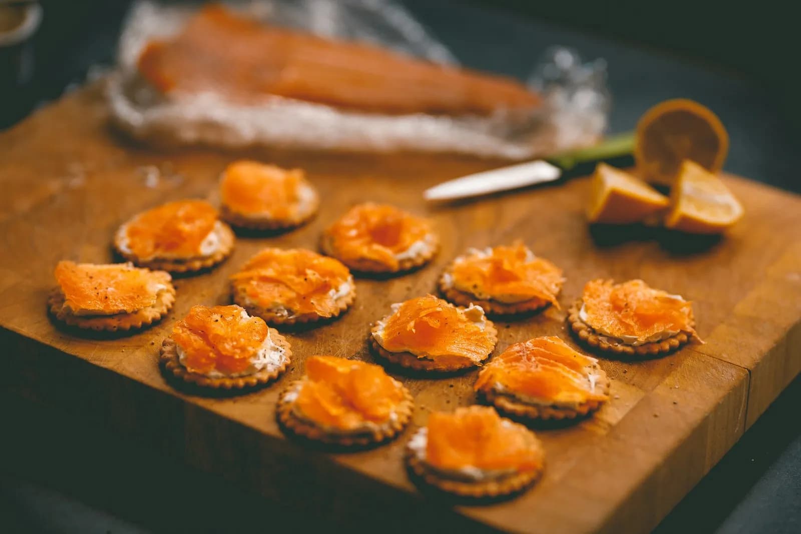 Canapés de salmón ahumado con queso crema sobre galletas, en una tabla de madera con limón y un lomo de salmón al fondo