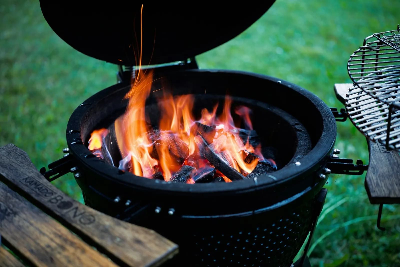 Kamado de cerámica negro con la tapa abierta y llamas vivas sobre el carbón, en un jardín al atardecer
