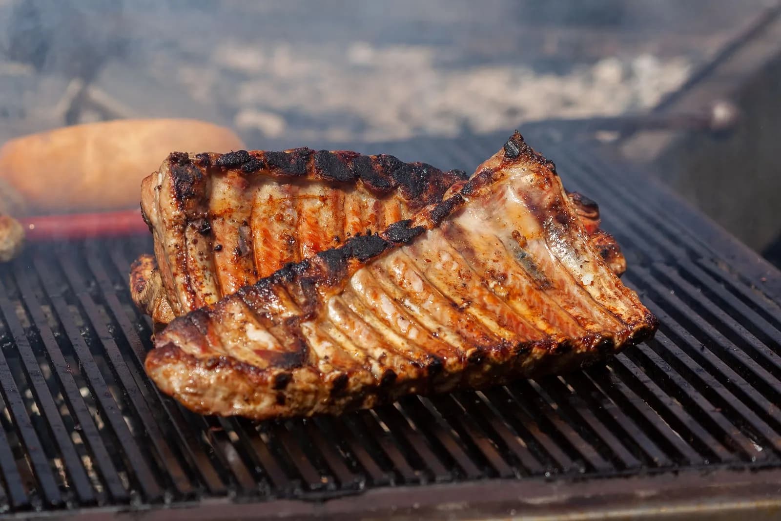 Dos costillares de cerdo dorados y churruscados por los bordes, sobre la parrilla de una barbacoa entre el humo