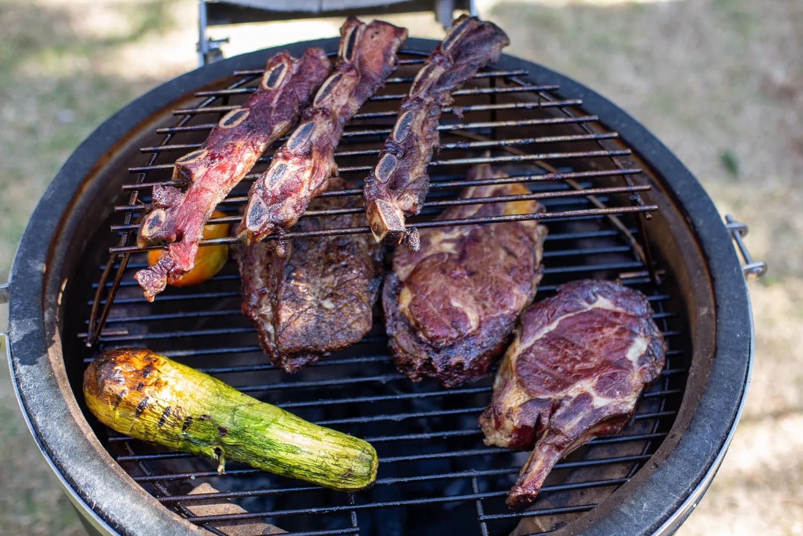 Kamado visto desde arriba, con costillas de vacuno sobre una rejilla elevada y chuletones y un calabacín en la parrilla