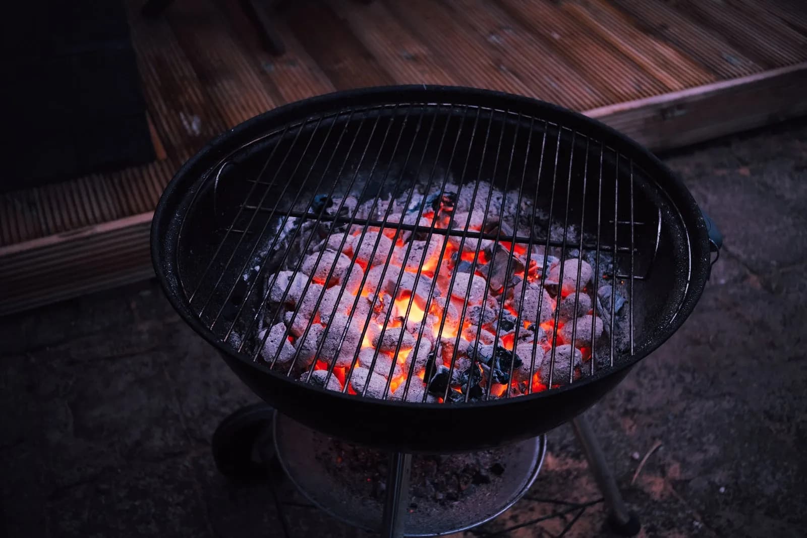 Kettle de carbón visto desde arriba, con las briquetas al rojo vivo bajo la parrilla, junto a una terraza de madera con poca luz