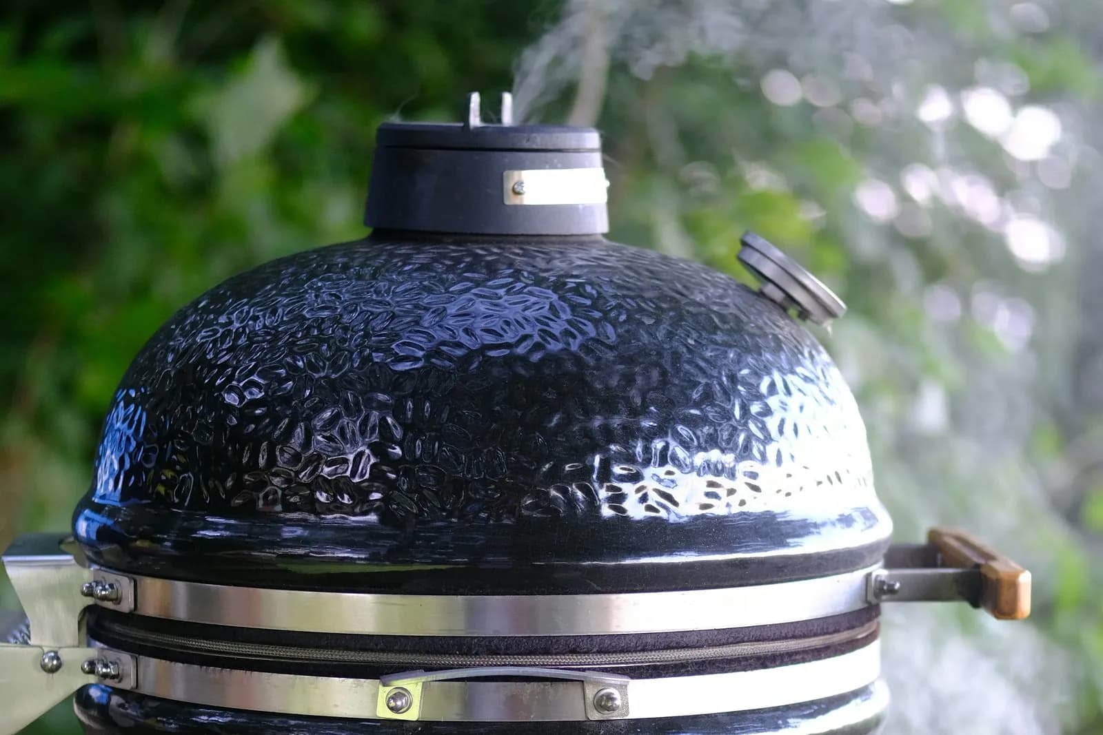 Cúpula de un kamado de cerámica negro y brillante, con un tenue hilo de humo junto a la chimenea y la junta entre las dos bandas de acero, ante follaje verde desenfocado
