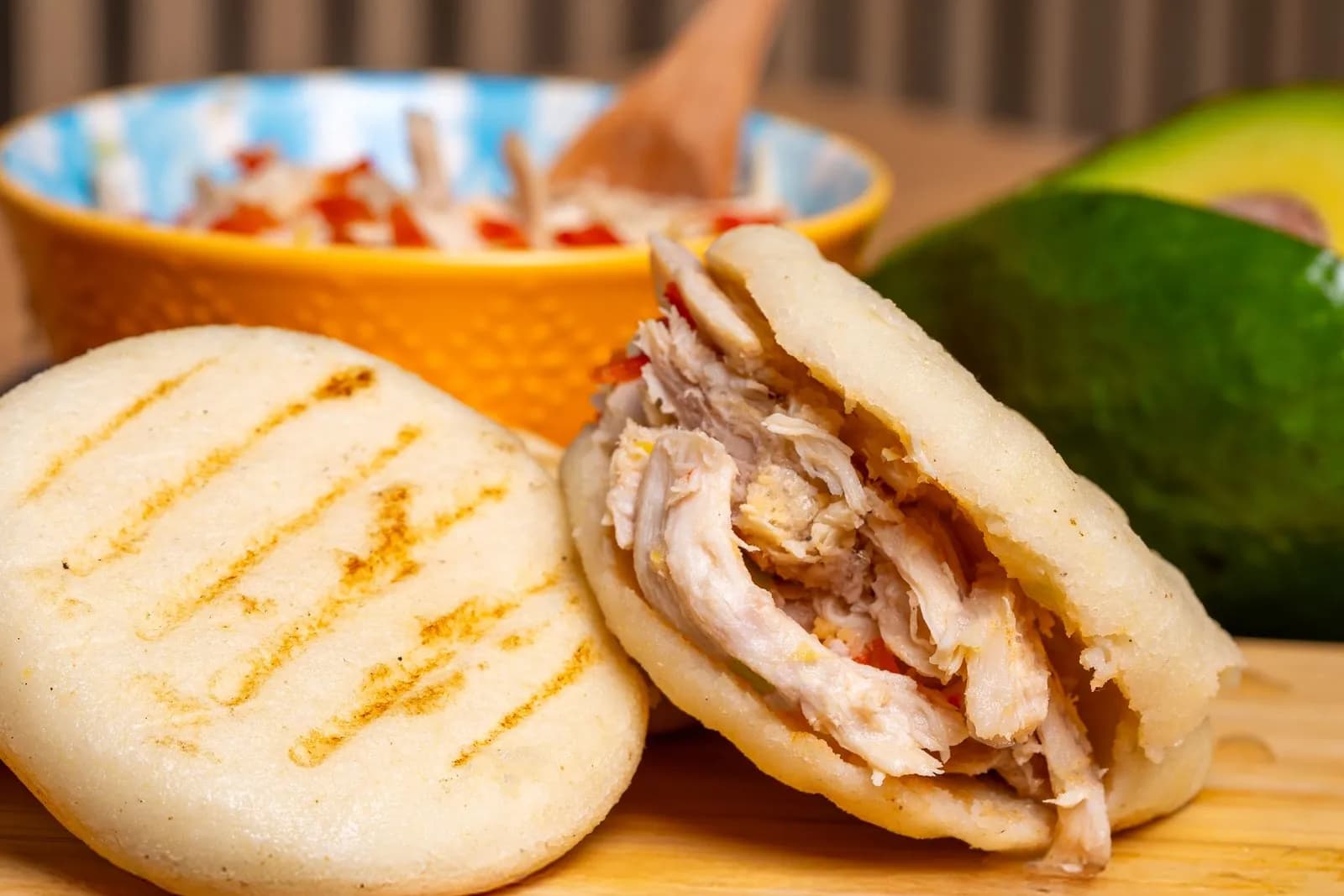 Dos arepas con rayas tostadas en la superficie: una entera y otra abierta por la mitad y rellena de pollo desmechado con trocitos de pimiento rojo; detrás, un cuenco naranja con más relleno y medio aguacate, sobre una tabla de madera clara