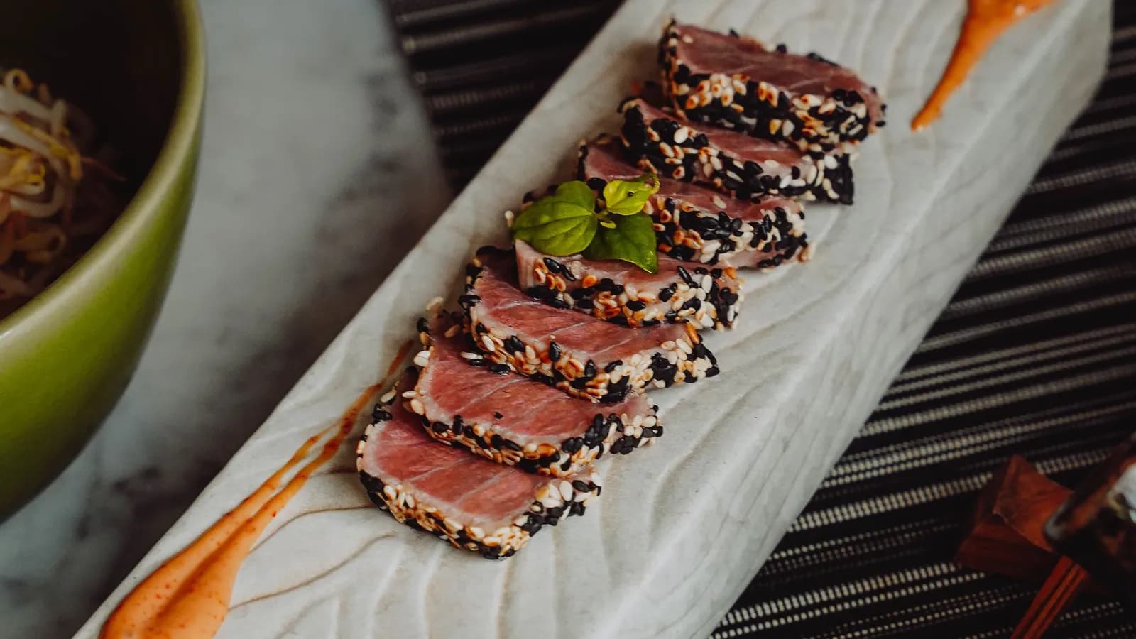 Lomo de atún rojo con costra de sésamo blanco y negro, sellado y cortado en láminas con el centro crudo y rojo, sobre una tabla de madera junto a un kamado cerámico encendido.