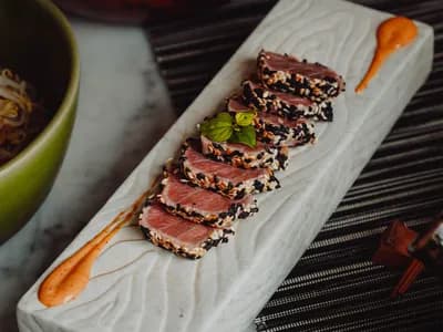 Lomo de atún rojo con costra de sésamo blanco y negro, sellado y cortado en láminas con el centro crudo y rojo, sobre una tabla de madera junto a un kamado cerámico encendido.