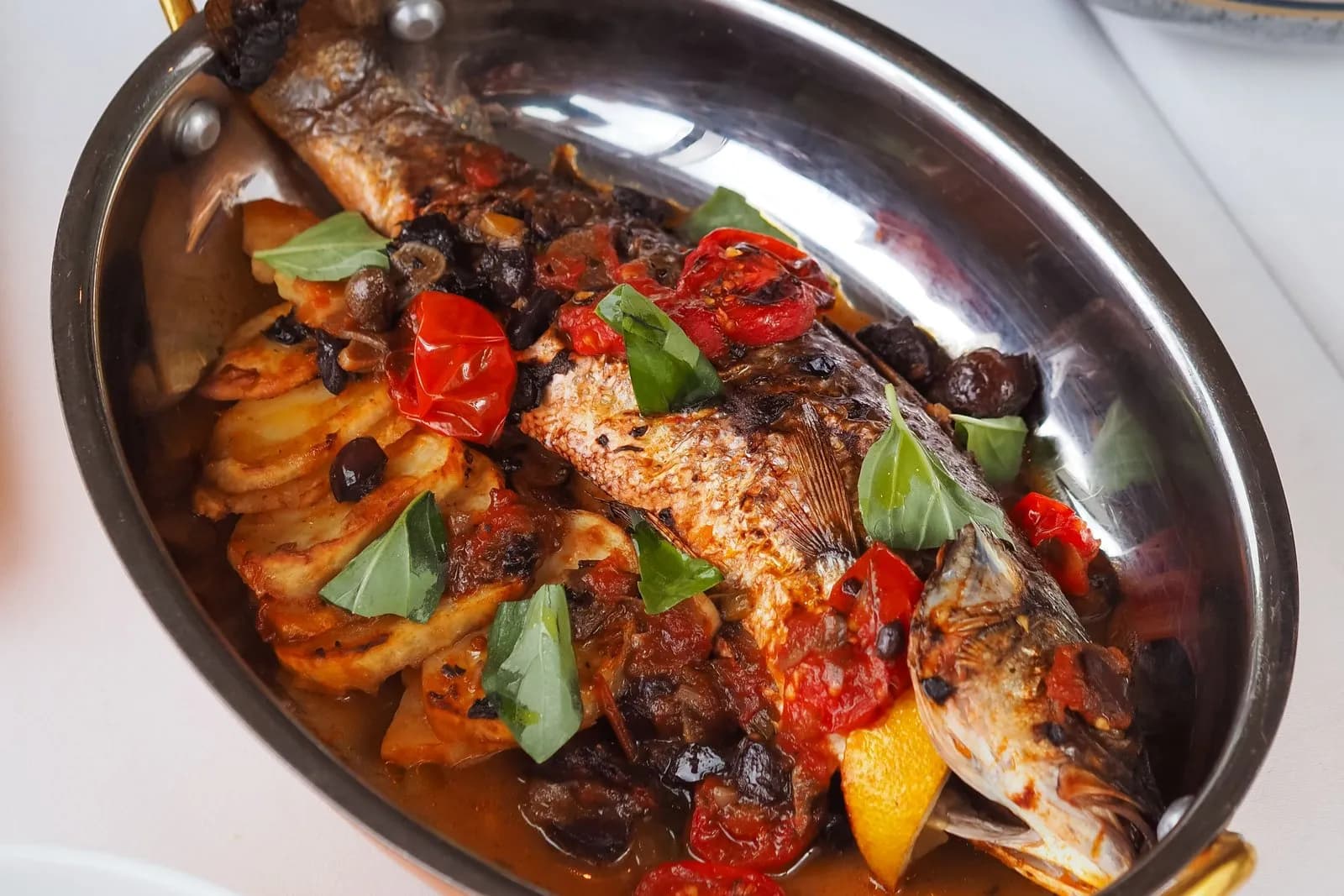 Pescado entero asado, de piel tostada, sobre una cama de rodajas de patata asadas en una fuente ovalada de cobre con asas doradas, con tomates cherry, aceitunas negras, hojas de albahaca y un gajo de limón