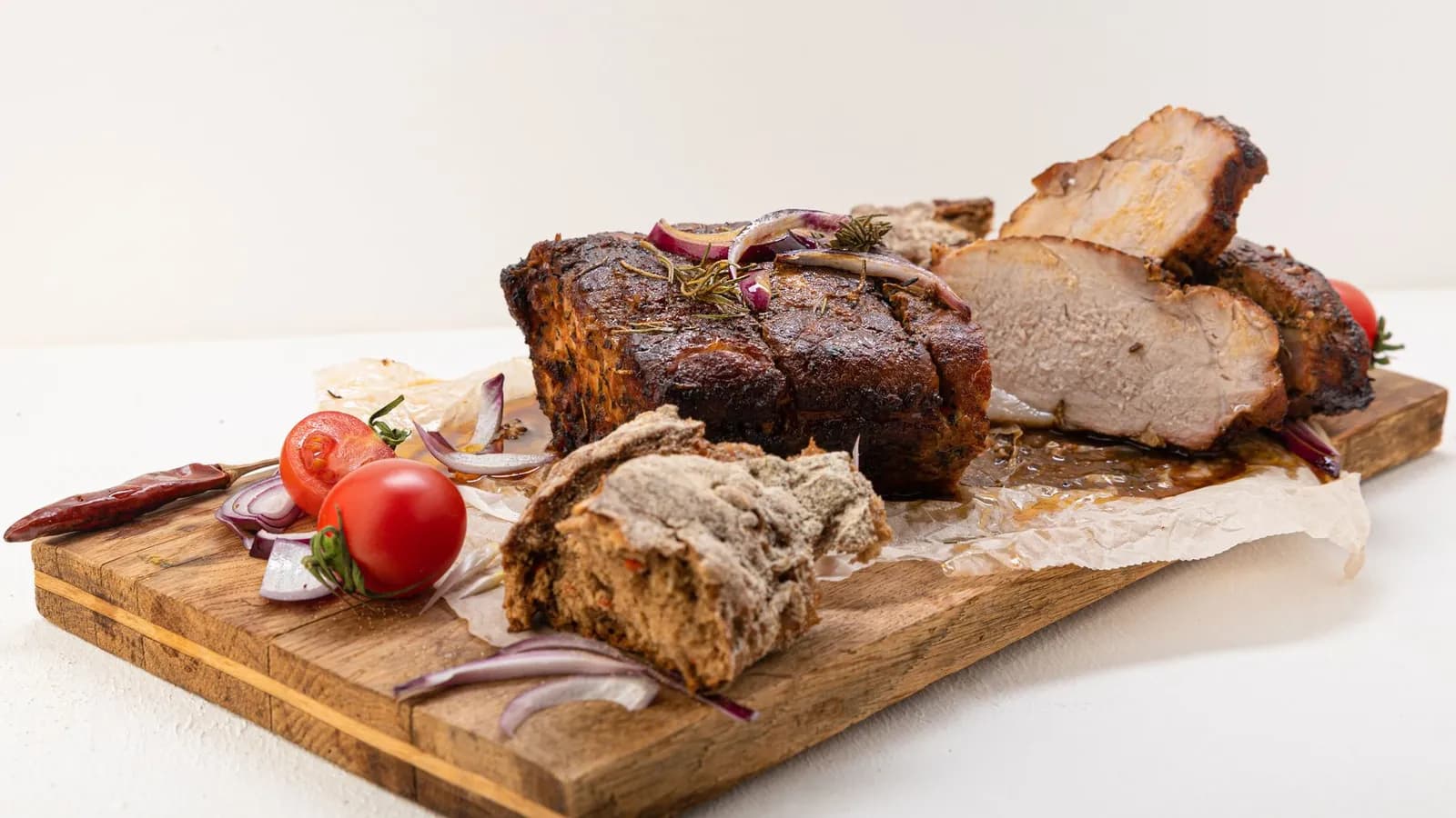 Asado de cerdo con costra oscura y romero, cortado en lonchas gruesas sobre tabla de madera con cebolla morada y tomates