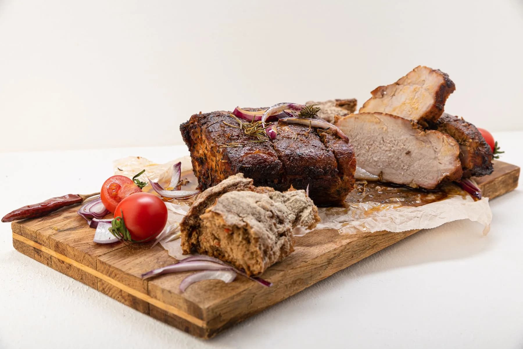 Asado de cerdo con costra oscura y romero, cortado en lonchas gruesas sobre tabla de madera con cebolla morada y tomates