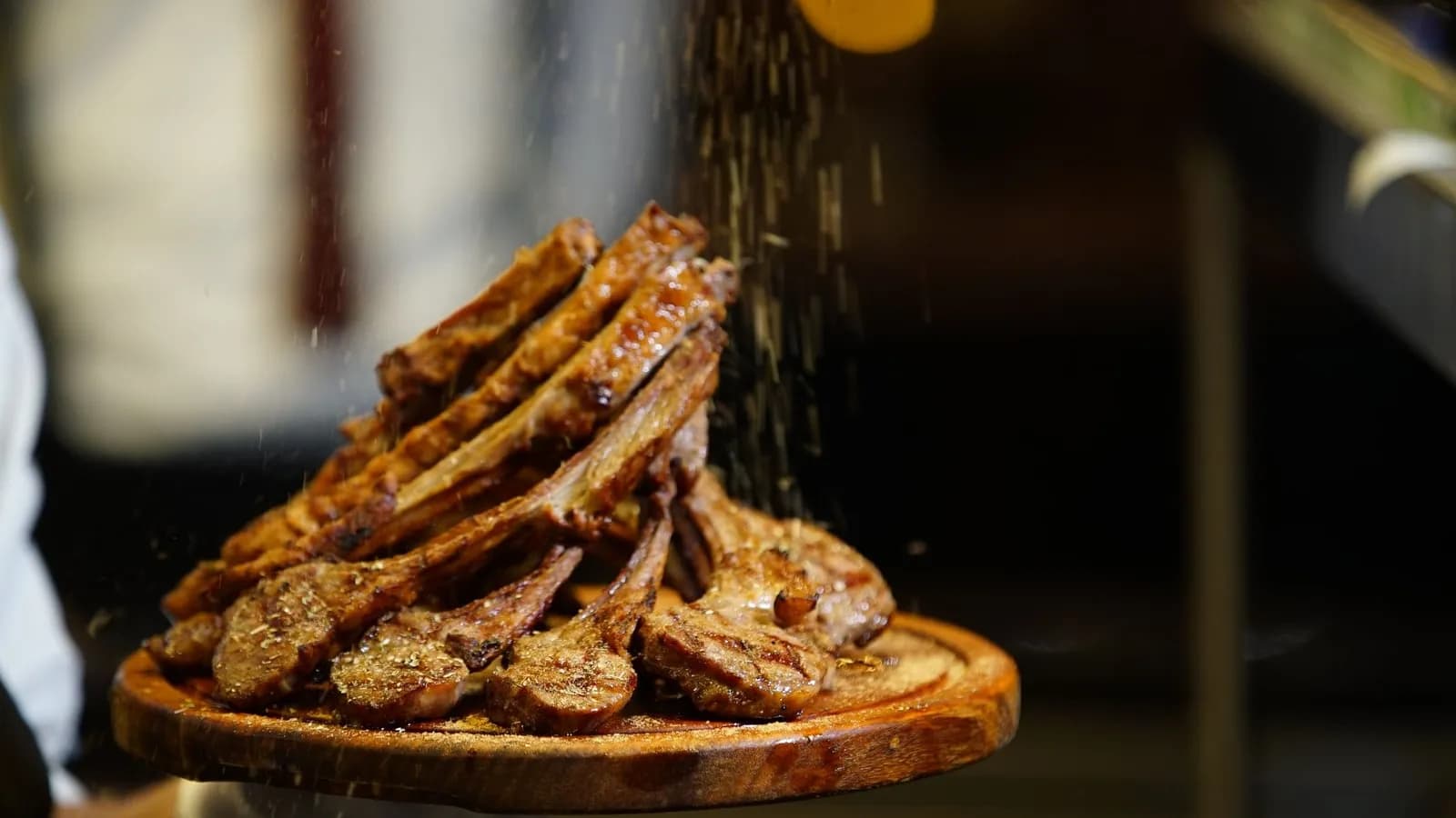 Pila de chuletillas de cordero a la parrilla, con marcas de rejilla en la carne y los huesos asomando, sobre una tabla redonda de madera que sostiene un cocinero, mientras cae por encima una lluvia de hierbas y especias secas, con un fondo oscuro desenfocado y un punto de luz anaranjado