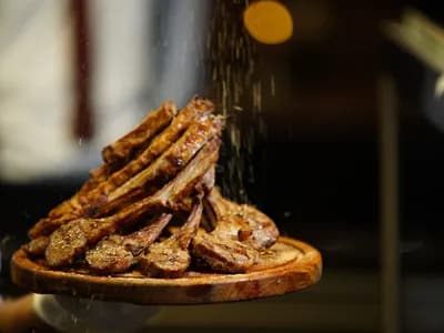 Pila de chuletillas de cordero a la parrilla, con marcas de rejilla en la carne y los huesos asomando, sobre una tabla redonda de madera que sostiene un cocinero, mientras cae por encima una lluvia de hierbas y especias secas, con un fondo oscuro desenfocado y un punto de luz anaranjado