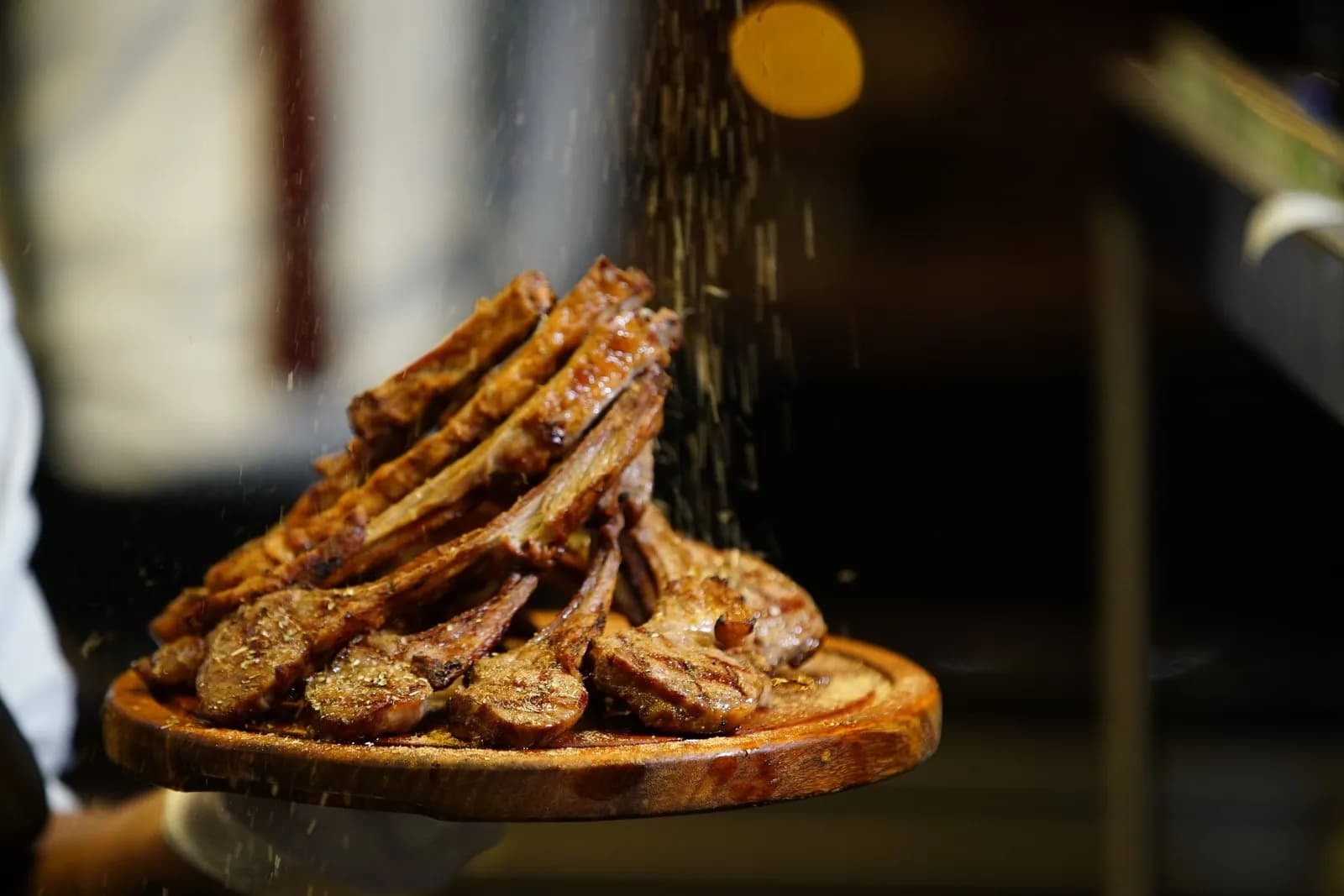 Pila de chuletillas de cordero a la parrilla, con marcas de rejilla en la carne y los huesos asomando, sobre una tabla redonda de madera que sostiene un cocinero, mientras cae por encima una lluvia de hierbas y especias secas, con un fondo oscuro desenfocado y un punto de luz anaranjado