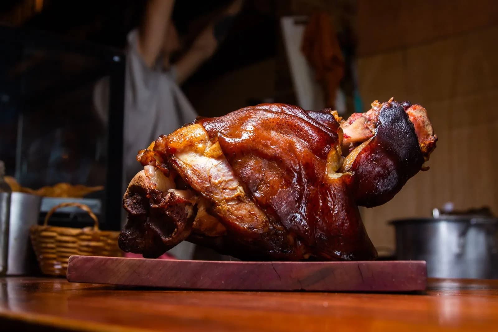 Codillo de cerdo entero asado sobre una tabla de madera, con la piel brillante de tono caoba y el hueso asomando por los extremos, ante una cocina desenfocada