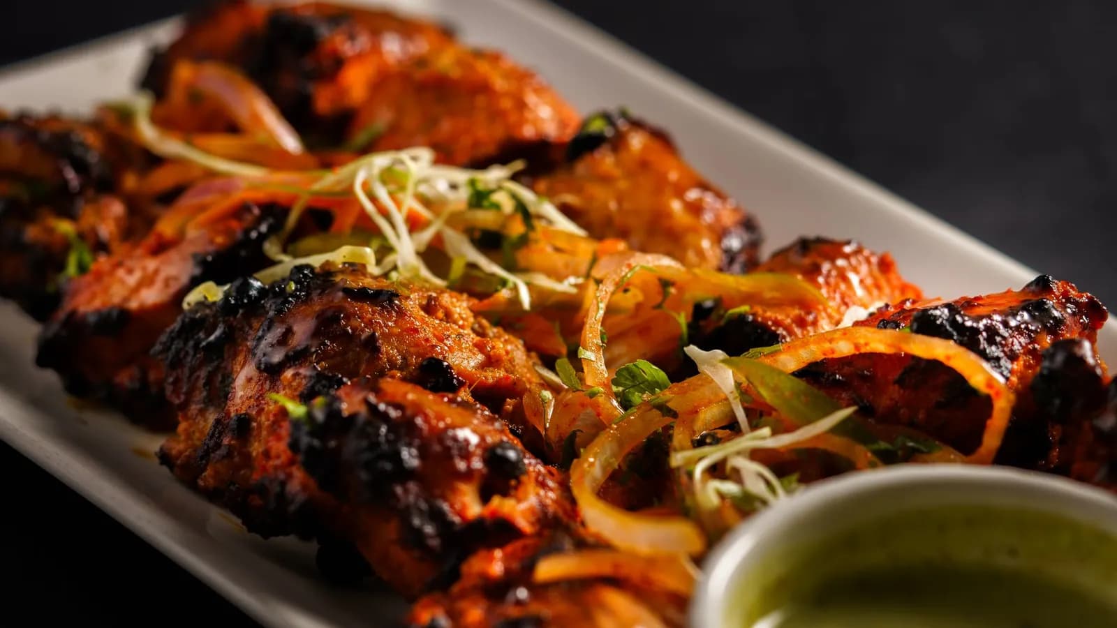 Contramuslos de pollo tandoori dorados y especiados sobre la parrilla de un kamado, junto a un cuenco de raita de pepino y gajos de lima.
