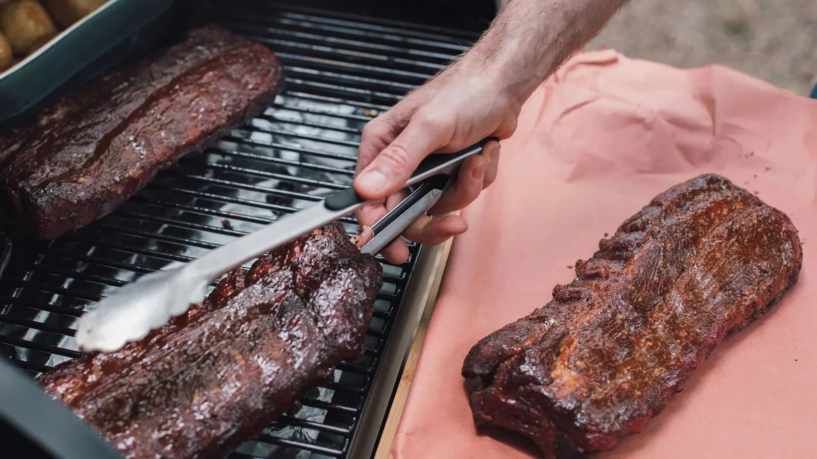 Tres costillares de cerdo con corteza oscura y brillante: dos sobre la rejilla de un ahumador, uno de ellos sujeto con unas pinzas, y un tercero reposando sobre papel de carnicero rosa