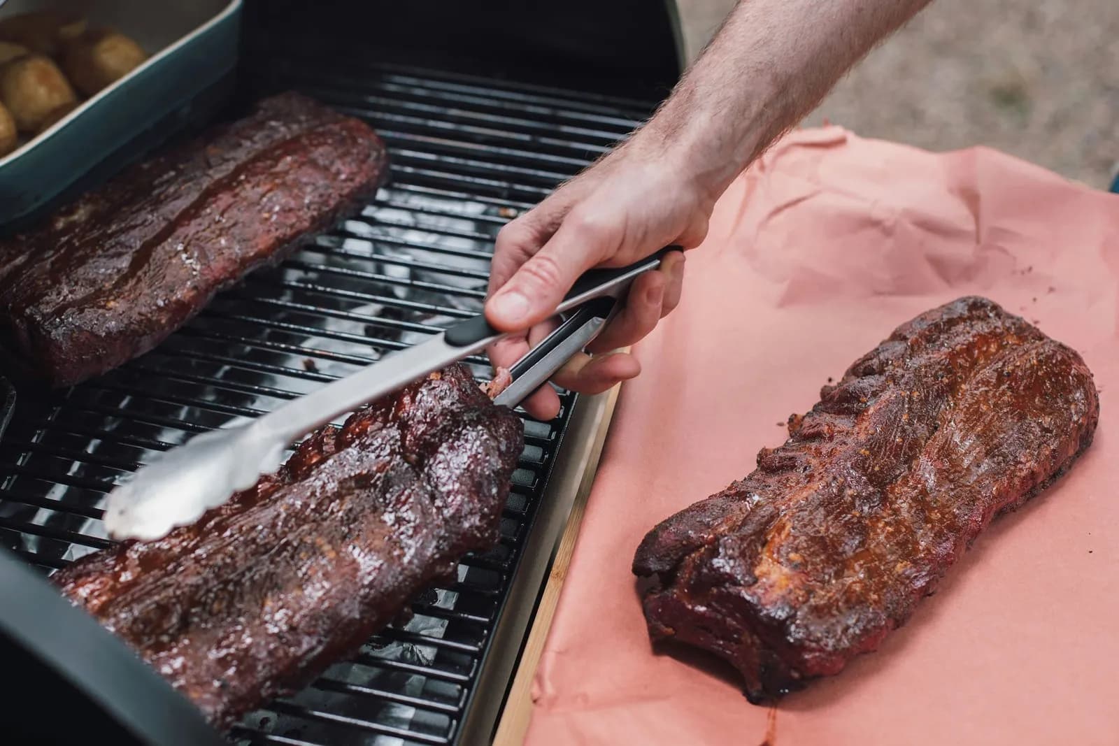 Tres costillares de cerdo con corteza oscura y brillante: dos sobre la rejilla de un ahumador, uno de ellos sujeto con unas pinzas, y un tercero reposando sobre papel de carnicero rosa