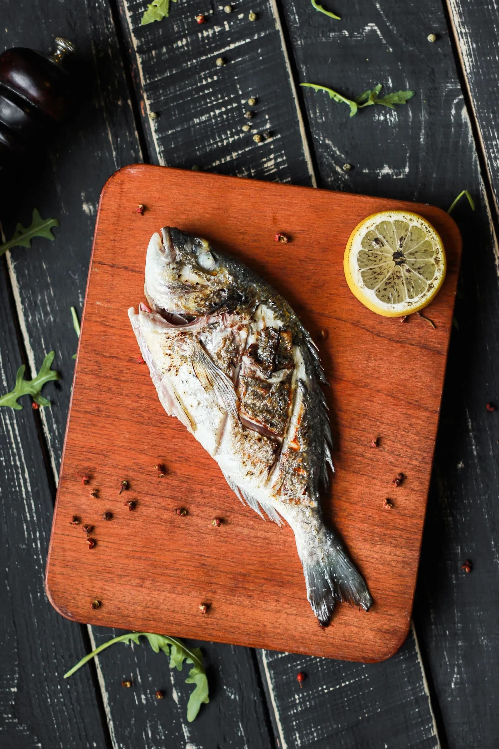 Dorada entera a la brasa con un limón asado y granos de pimienta sobre una tabla de madera oscura