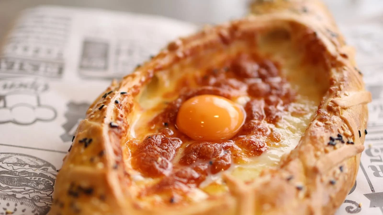 Jachapuri adjaruli con la masa dorada y salpicada de semillas, el queso fundido y gratinado en el centro y una yema entera encima, sobre papel impreso