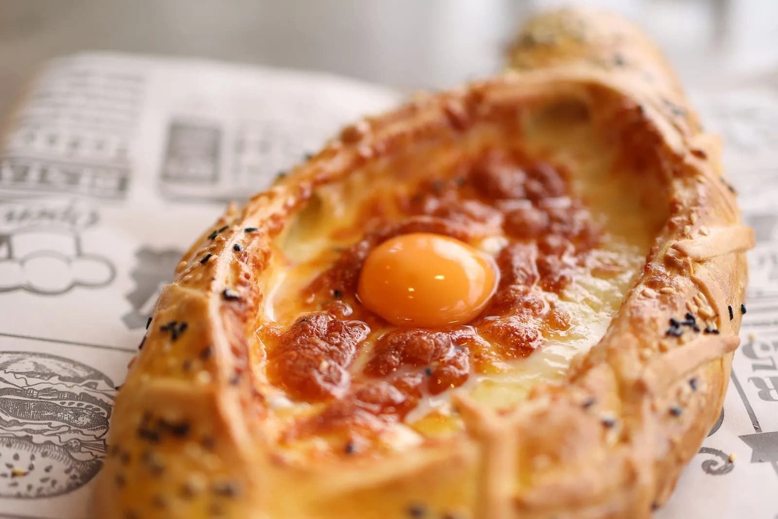 Jachapuri adjaruli con la masa dorada y salpicada de semillas, el queso fundido y gratinado en el centro y una yema entera encima, sobre papel impreso