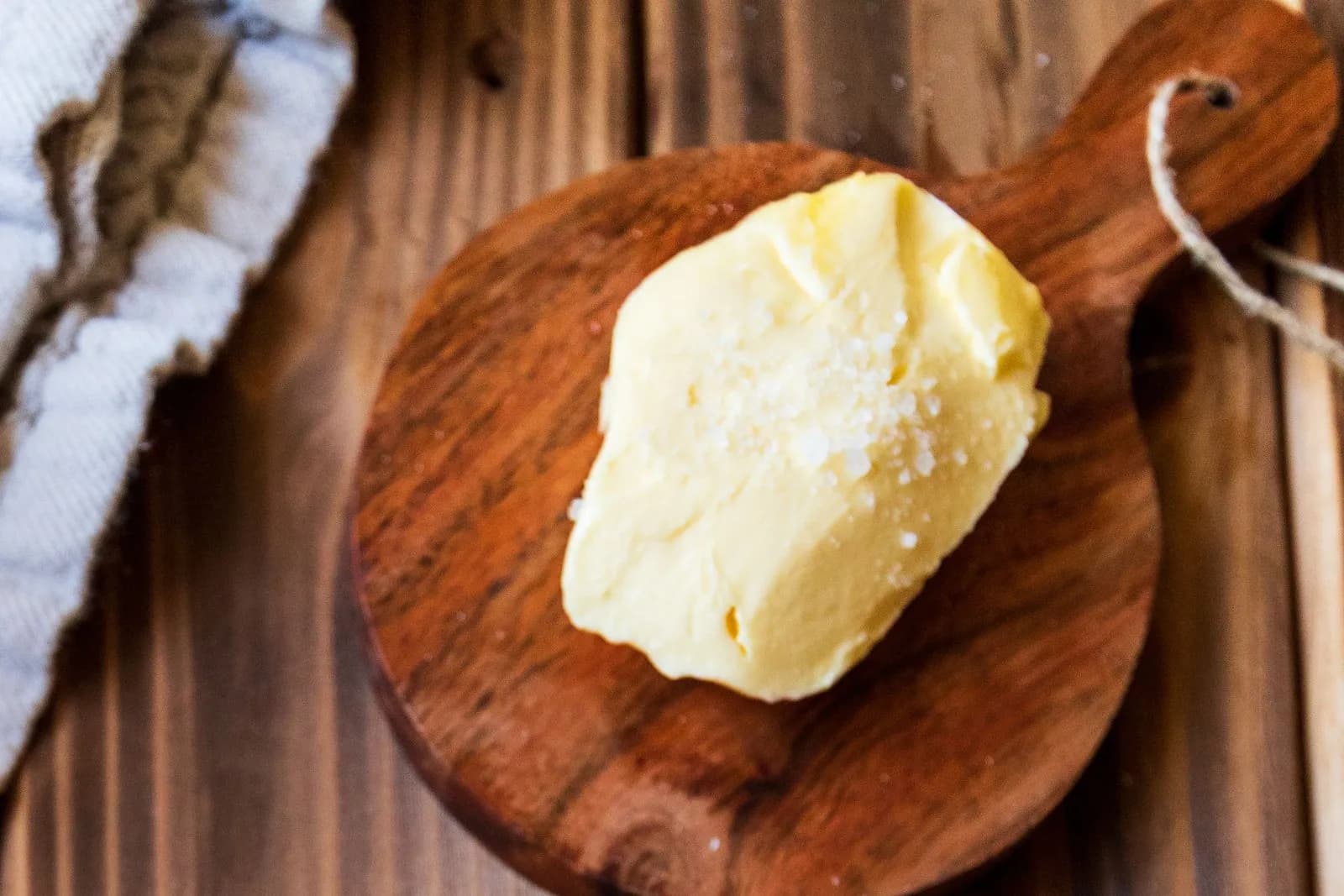 Porción de mantequilla cremosa y dorada con escamas de sal gruesa por encima, sobre una pequeña tabla de madera redonda con mango y cordel, en una mesa de tablones oscuros junto a un paño blanco