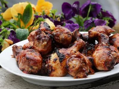 Montón de muslos de pollo con un glaseado oscuro y brillante y zonas churruscadas, servidos en un plato blanco sobre madera envejecida, con flores amarillas y moradas desenfocadas al fondo