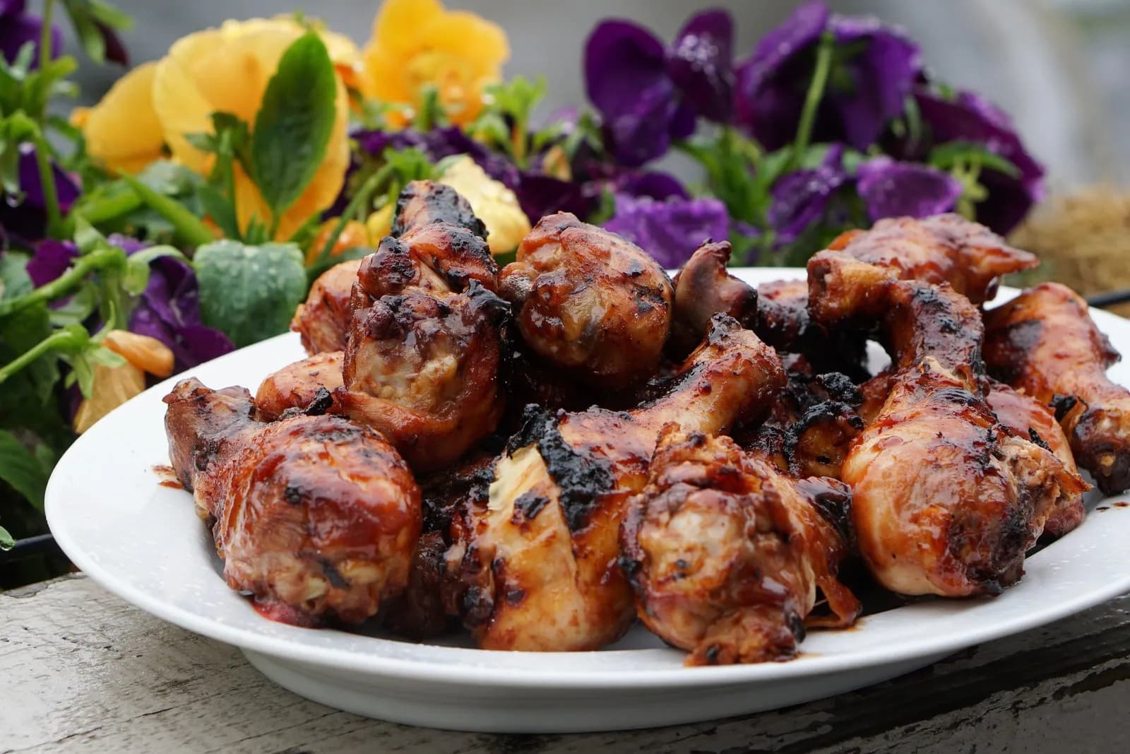 Montón de muslos de pollo con un glaseado oscuro y brillante y zonas churruscadas, servidos en un plato blanco sobre madera envejecida, con flores amarillas y moradas desenfocadas al fondo