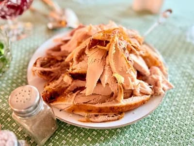 Plato blanco con un montón de pavo asado en trozos y lonchas, carne blanca con la piel dorada, sobre un mantel verde; en primer plano un salero y un pimentero de cristal y, desenfocados, cuencos con salsa de arándanos y brócoli