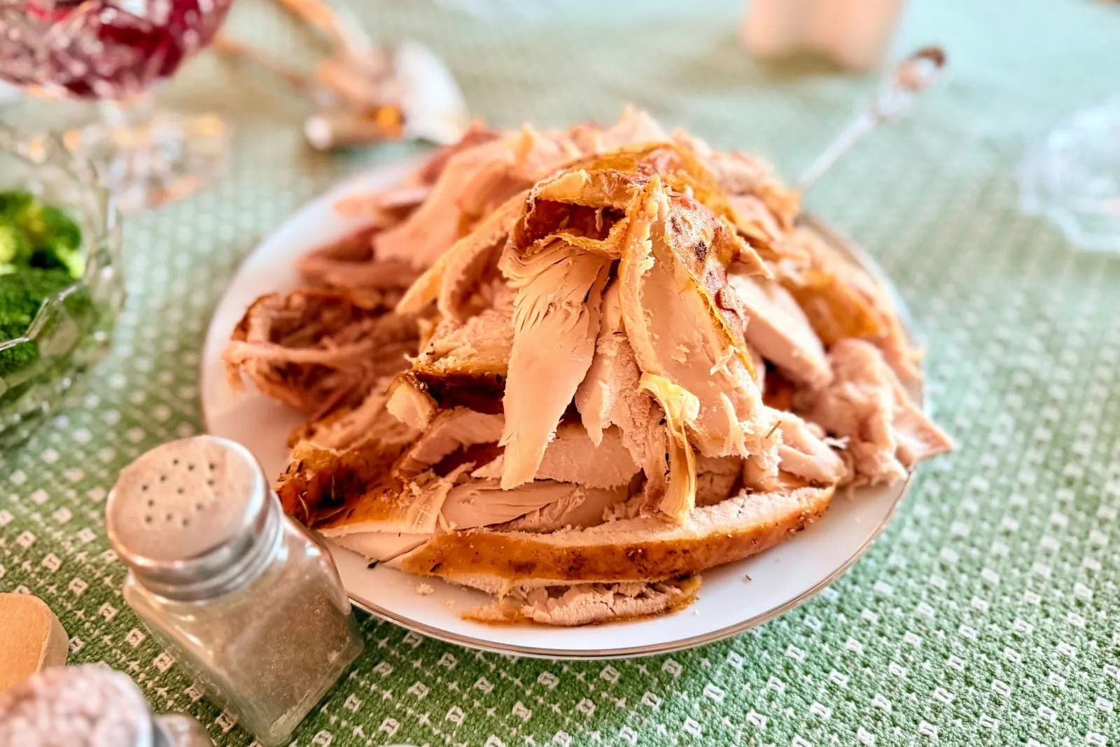 Plato blanco con un montón de pavo asado en trozos y lonchas, carne blanca con la piel dorada, sobre un mantel verde; en primer plano un salero y un pimentero de cristal y, desenfocados, cuencos con salsa de arándanos y brócoli