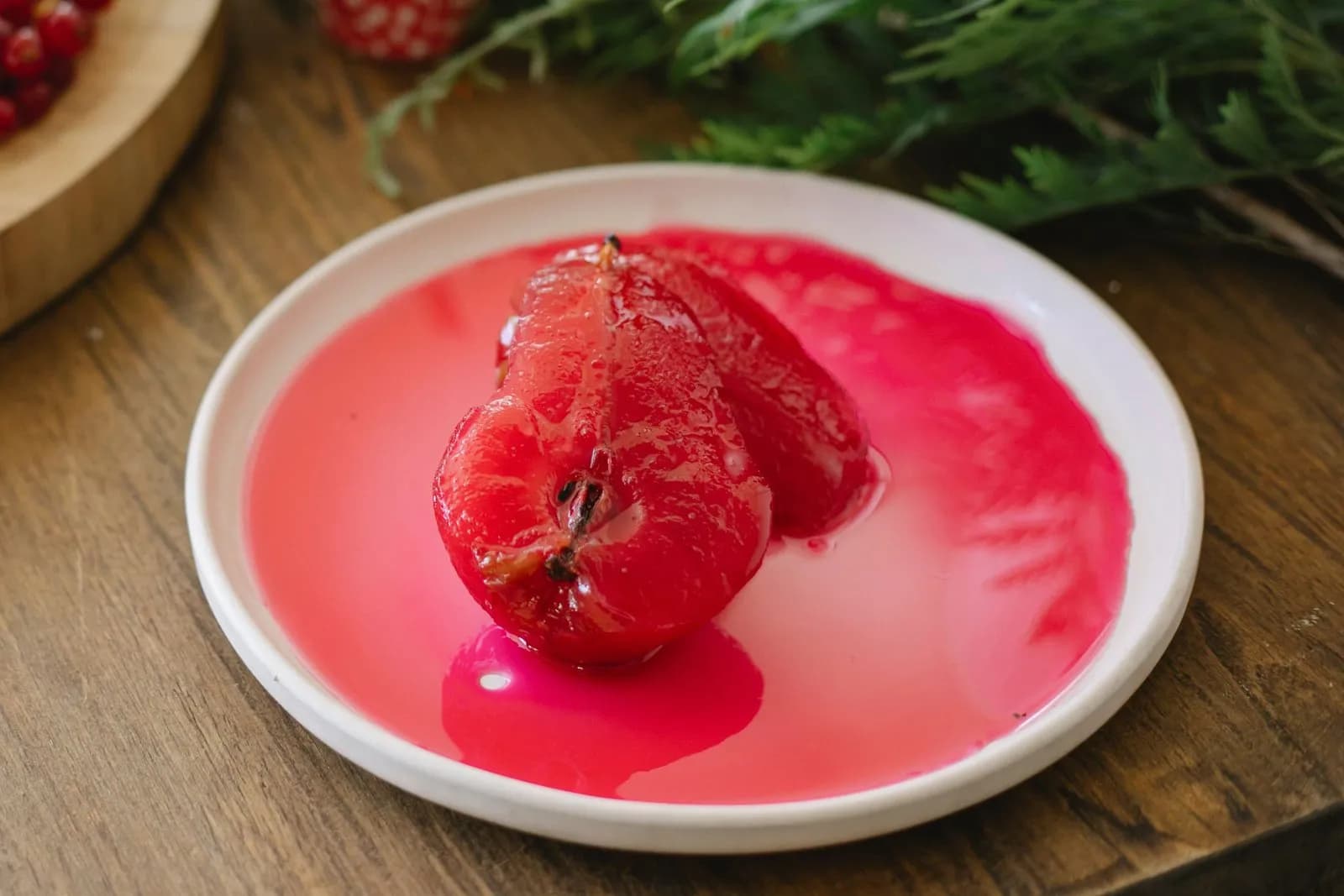 Una pera pochada partida en dos mitades, de un rojo intenso y brillante, en almíbar rosado sobre un plato blanco; al fondo, un bizcocho con grosellas y ramas de abeto