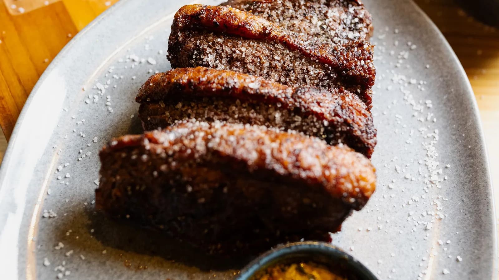 Slices of chargrilled beef with a dark crust on a grey plate, next to a bowl of chimichurri
