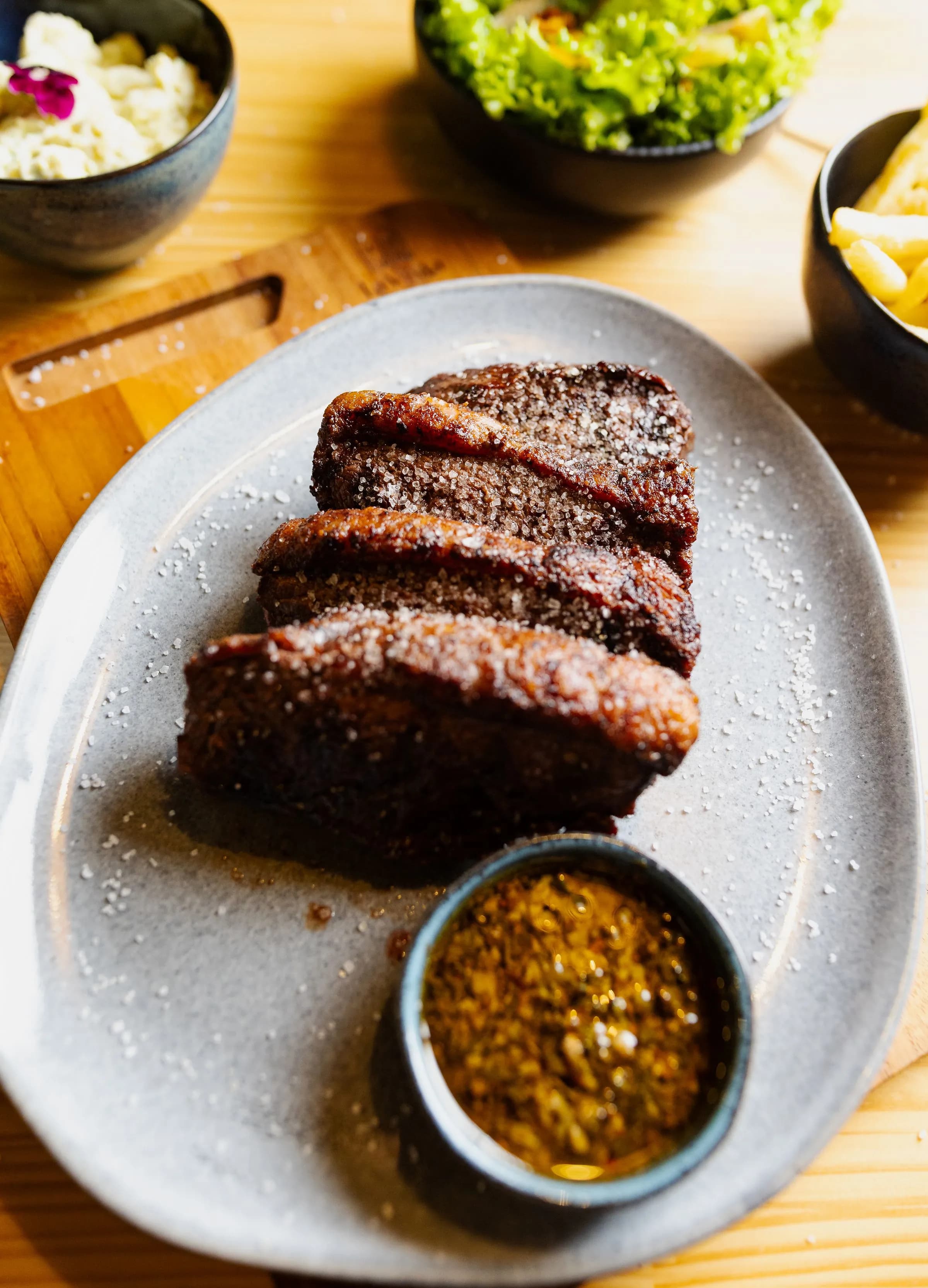 Lonchas de carne a la brasa con costra oscura en un plato gris, junto a un cuenco de chimichurri