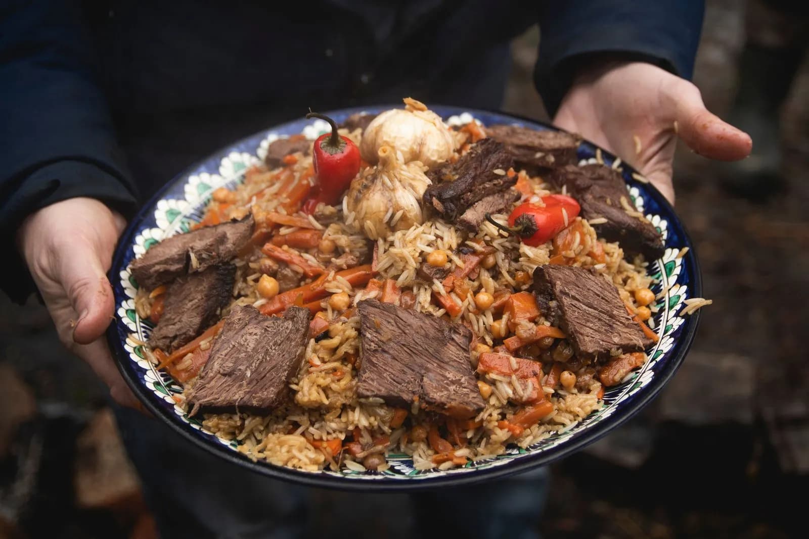 Plov uzbeko servido en una fuente de cerámica con el borde decorado en azul, blanco y verde que una persona sostiene con las dos manos al aire libre: arroz suelto con tiras gruesas de zanahoria y garbanzos, grandes lonchas de carne por encima, dos cabezas de ajo enteras y dos guindillas rojas (versión con garbanzos)