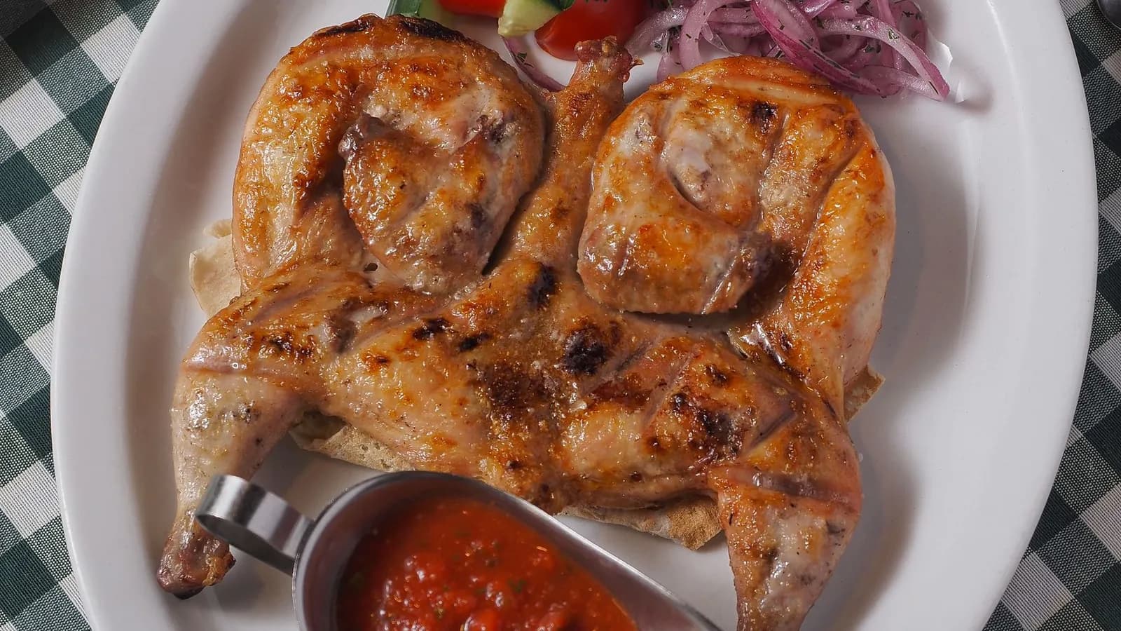 Picantón aplanado a la tabaka con piel dorada, salsa de tomate, cebolla encurtida y pan sobre plato blanco
