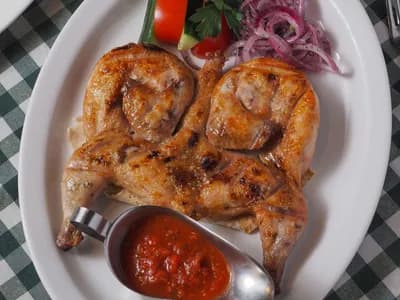 Picantón aplanado a la tabaka con piel dorada, salsa de tomate, cebolla encurtida y pan sobre plato blanco