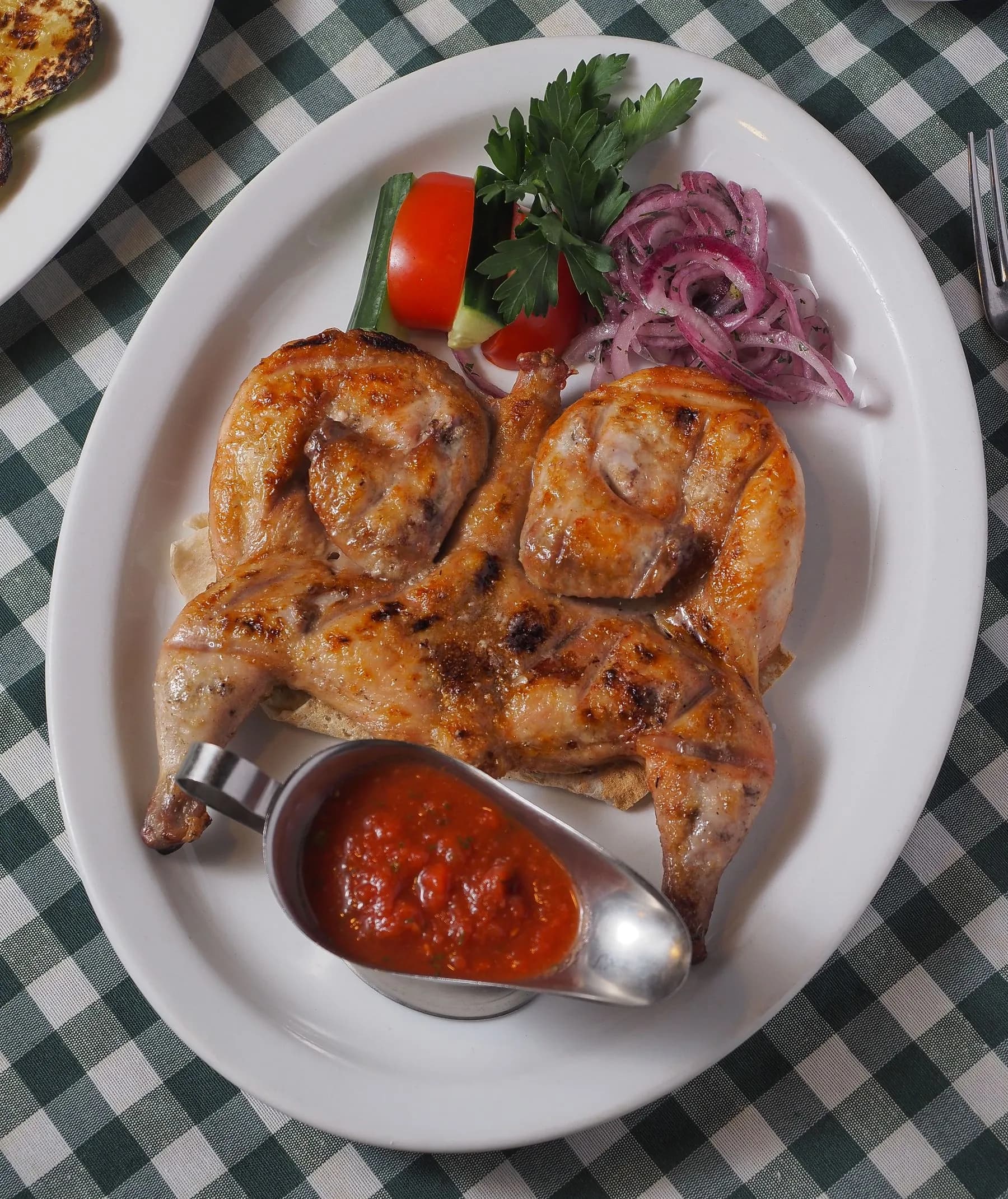 Picantón aplanado a la tabaka con piel dorada, salsa de tomate, cebolla encurtida y pan sobre plato blanco