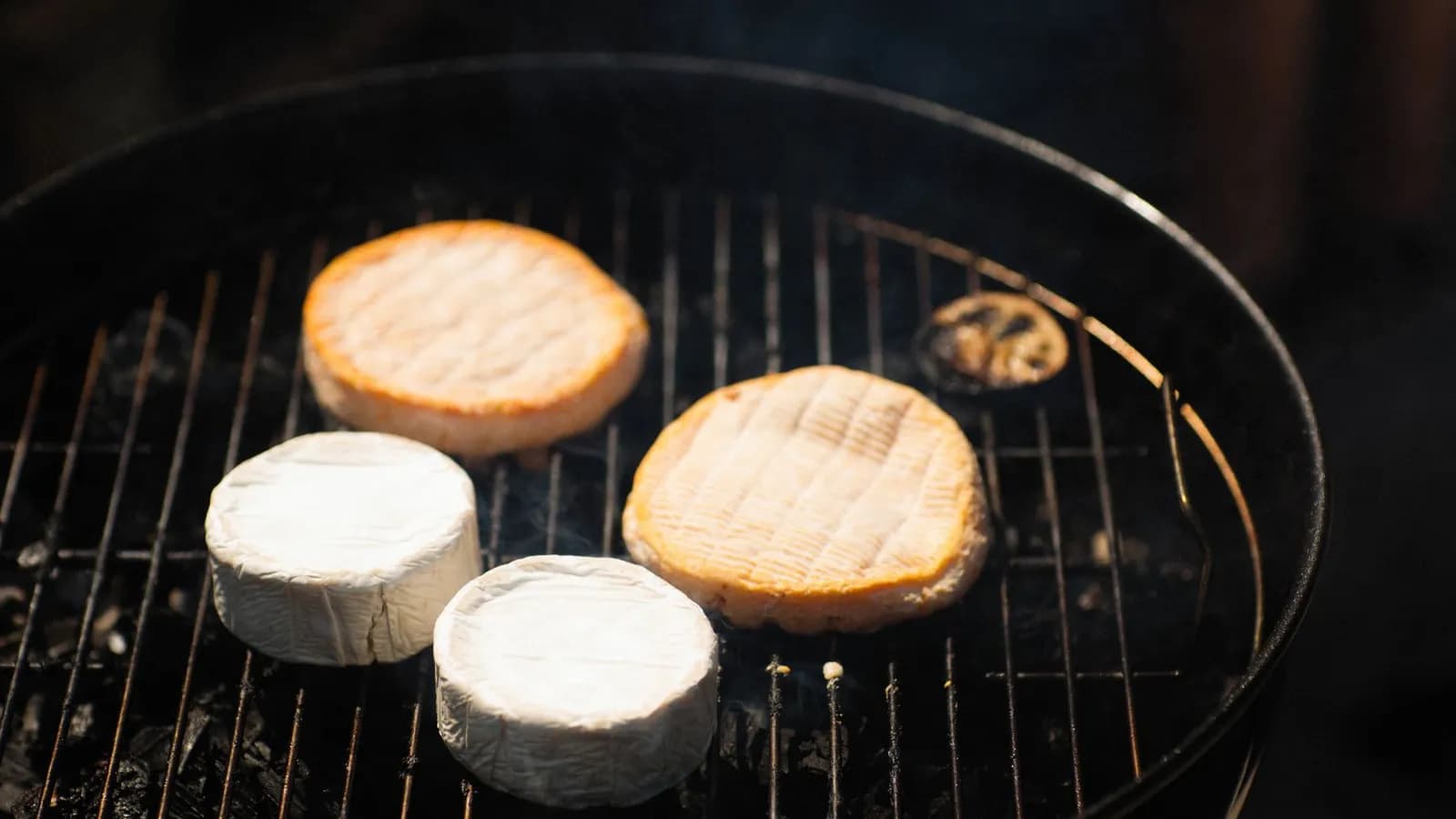 Cuatro quesos redondos ahumándose sobre la parrilla de una barbacoa negra con humo tenue
