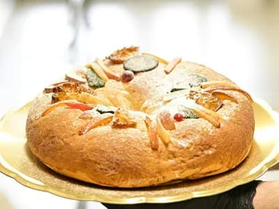 Rosca de Reyes de estilo mexicano entera, sobre una bandeja dorada de borde festoneado que sostienen unas manos, una con guante negro: corona de masa dorada espolvoreada de azúcar glas y cubierta de fruta confitada de colores (rodajas caramelizadas, tiras anaranjadas y piezas verdes, rojas y oscuras), ante un fondo claro desenfocado