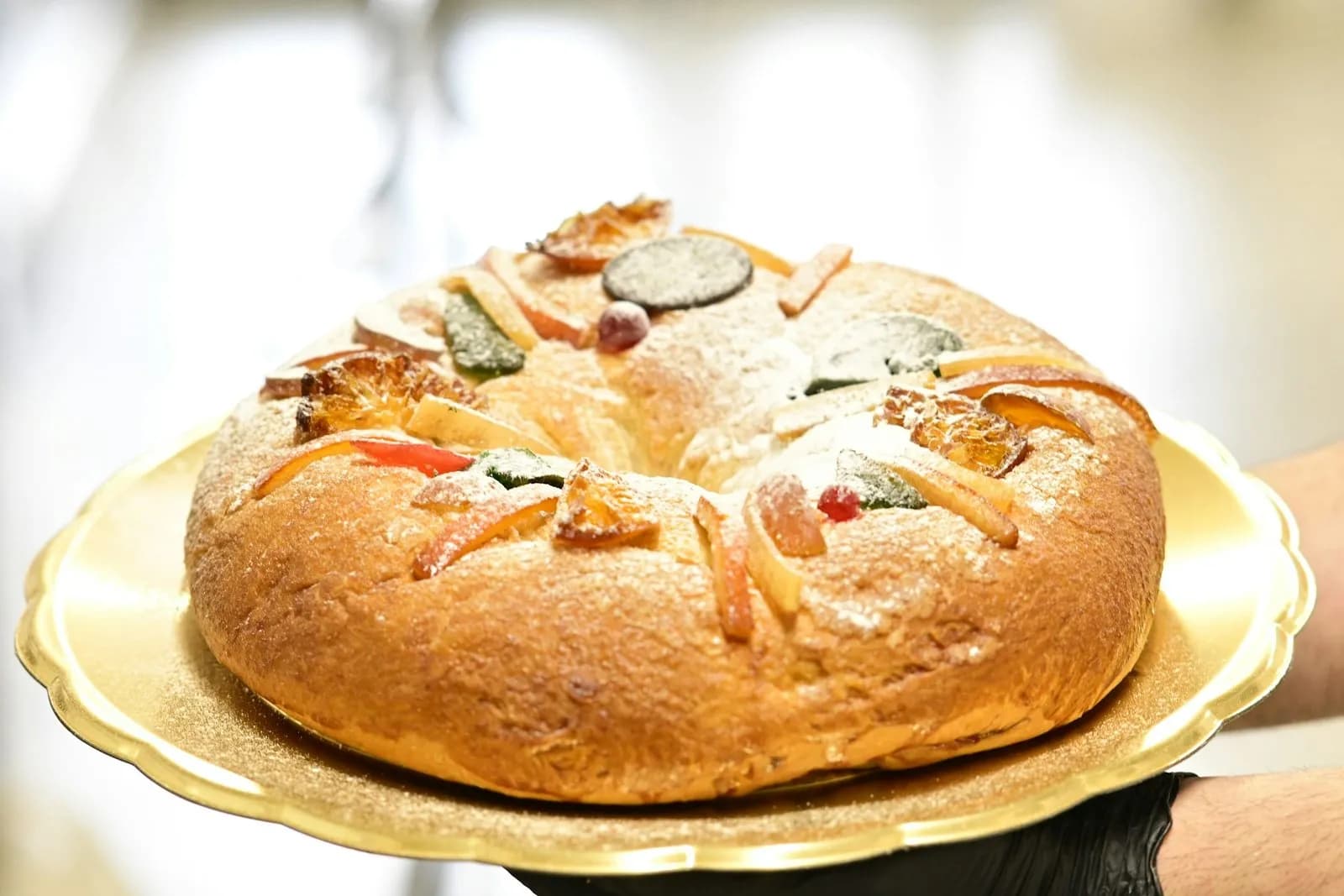 Rosca de Reyes de estilo mexicano entera, sobre una bandeja dorada de borde festoneado que sostienen unas manos, una con guante negro: corona de masa dorada espolvoreada de azúcar glas y cubierta de fruta confitada de colores (rodajas caramelizadas, tiras anaranjadas y piezas verdes, rojas y oscuras), ante un fondo claro desenfocado