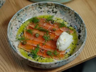 Lonchas finas de salmón ahumado con eneldo, alcaparras y sésamo en un cuenco de cerámica moteada, con aceite de oliva y una cucharada de crema blanca, sobre una mesa de madera clara
