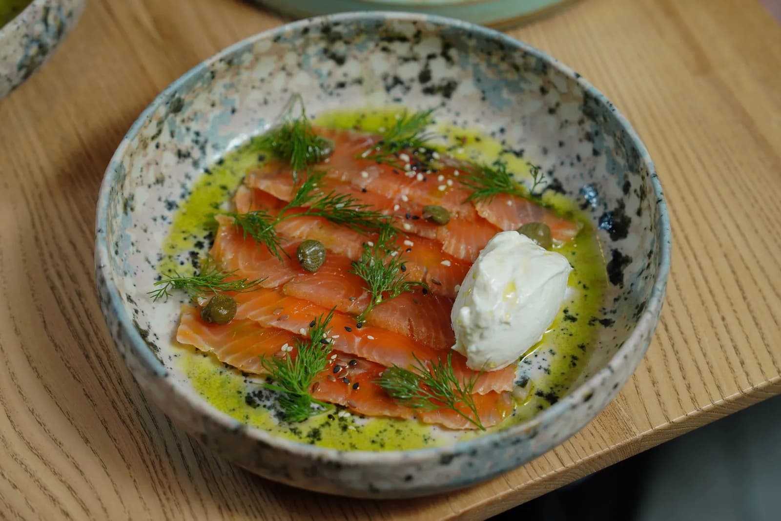 Lonchas finas de salmón ahumado con eneldo, alcaparras y sésamo en un cuenco de cerámica moteada, con aceite de oliva y una cucharada de crema blanca, sobre una mesa de madera clara