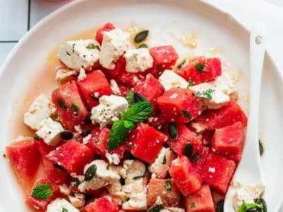 Triángulos de sandía con marcas de parrilla, feta desmenuzado, hojas de menta e hilos de reducción de balsámico sobre una tabla de roble junto a un kamado encendido.