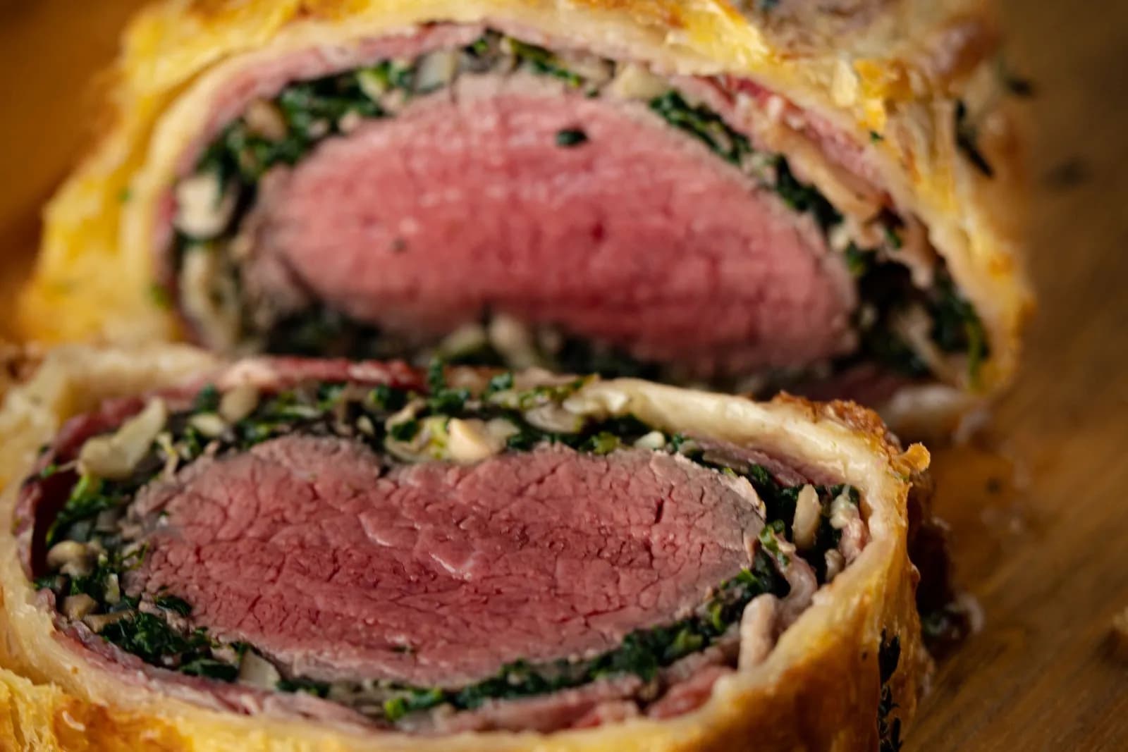 Primer plano de un solomillo Wellington cortado: el centro de vacuno rosado, rodeado de una capa fina de jamón, otra de espinacas y champiñón picados y hojaldre dorado, con una rodaja separada delante sobre una tabla de madera