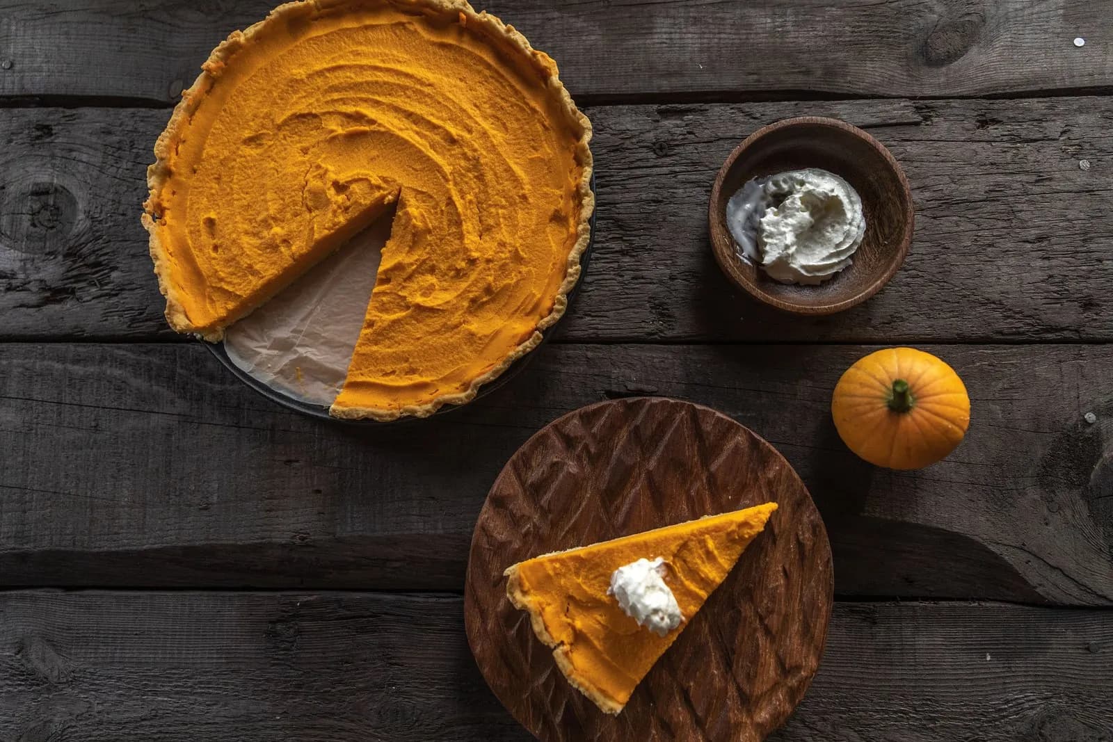 Tarta de calabaza vista desde arriba, de relleno naranja liso y borde de masa dorada, con una porción retirada; debajo, una porción con nata montada sobre una tabla de madera tallada, y a la derecha un cuenco de nata y una calabaza decorativa pequeña, sobre tablones oscuros