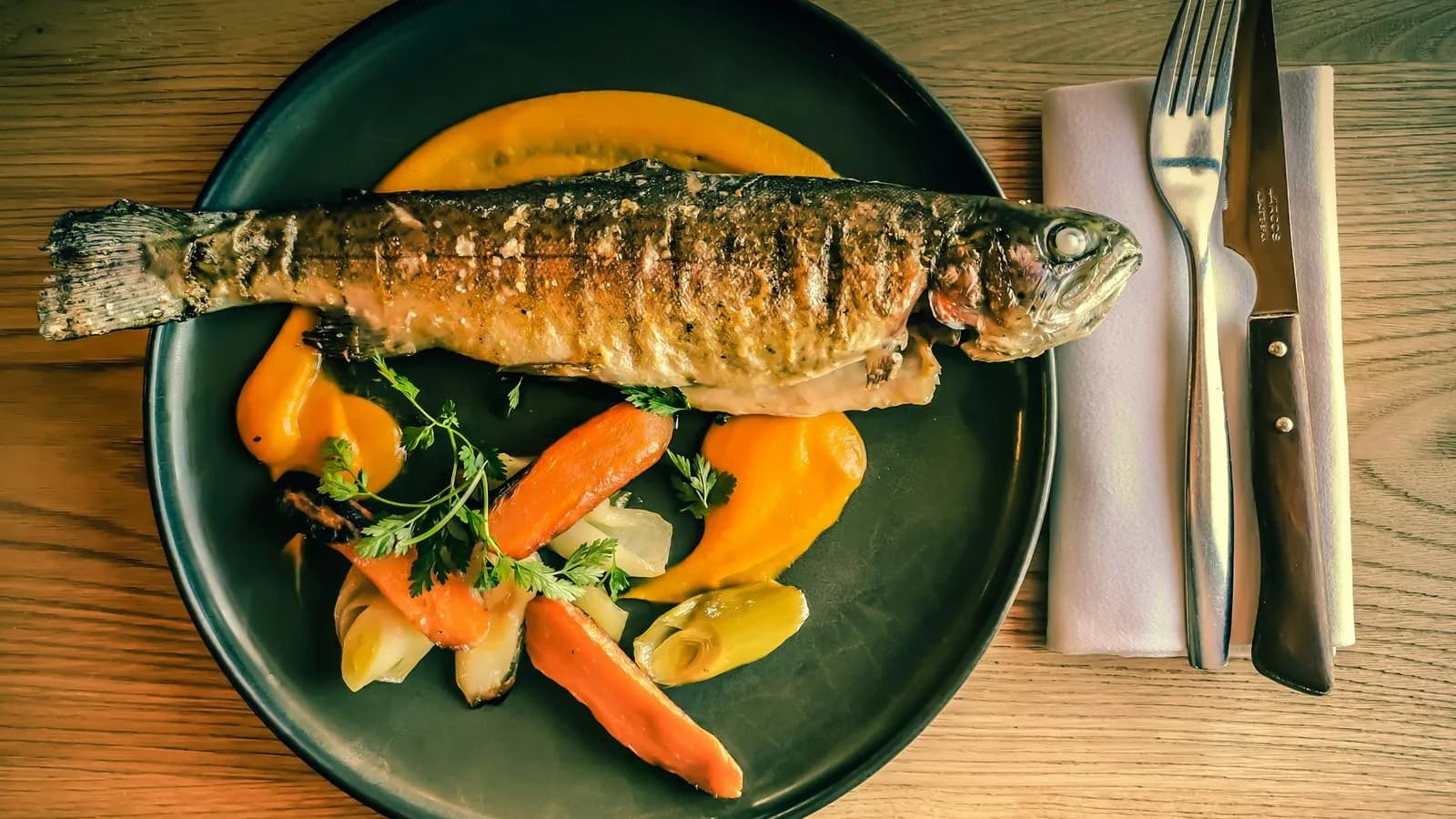 Trucha entera de piel dorada, marcada por los barrotes de la parrilla y salpicada de sal en escamas, en un plato oscuro con zanahorias asadas, puerro, perifollo y puré anaranjado, junto a un tenedor y un cuchillo sobre una servilleta