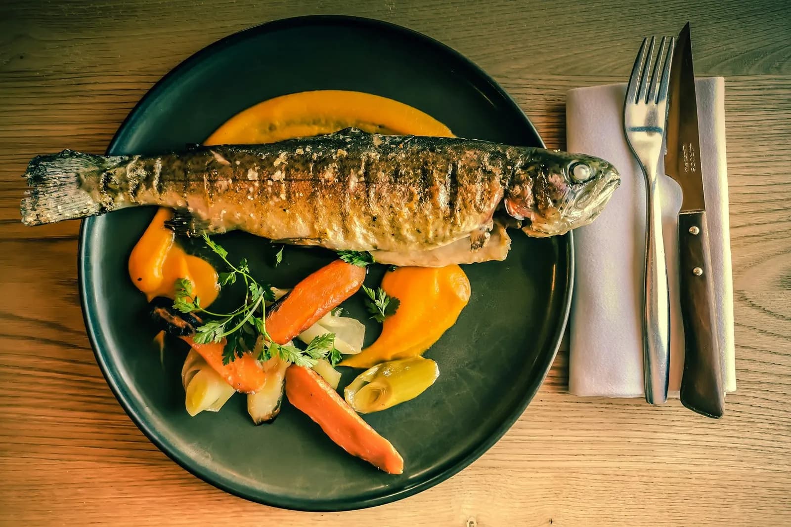 Trucha entera de piel dorada, marcada por los barrotes de la parrilla y salpicada de sal en escamas, en un plato oscuro con zanahorias asadas, puerro, perifollo y puré anaranjado, junto a un tenedor y un cuchillo sobre una servilleta