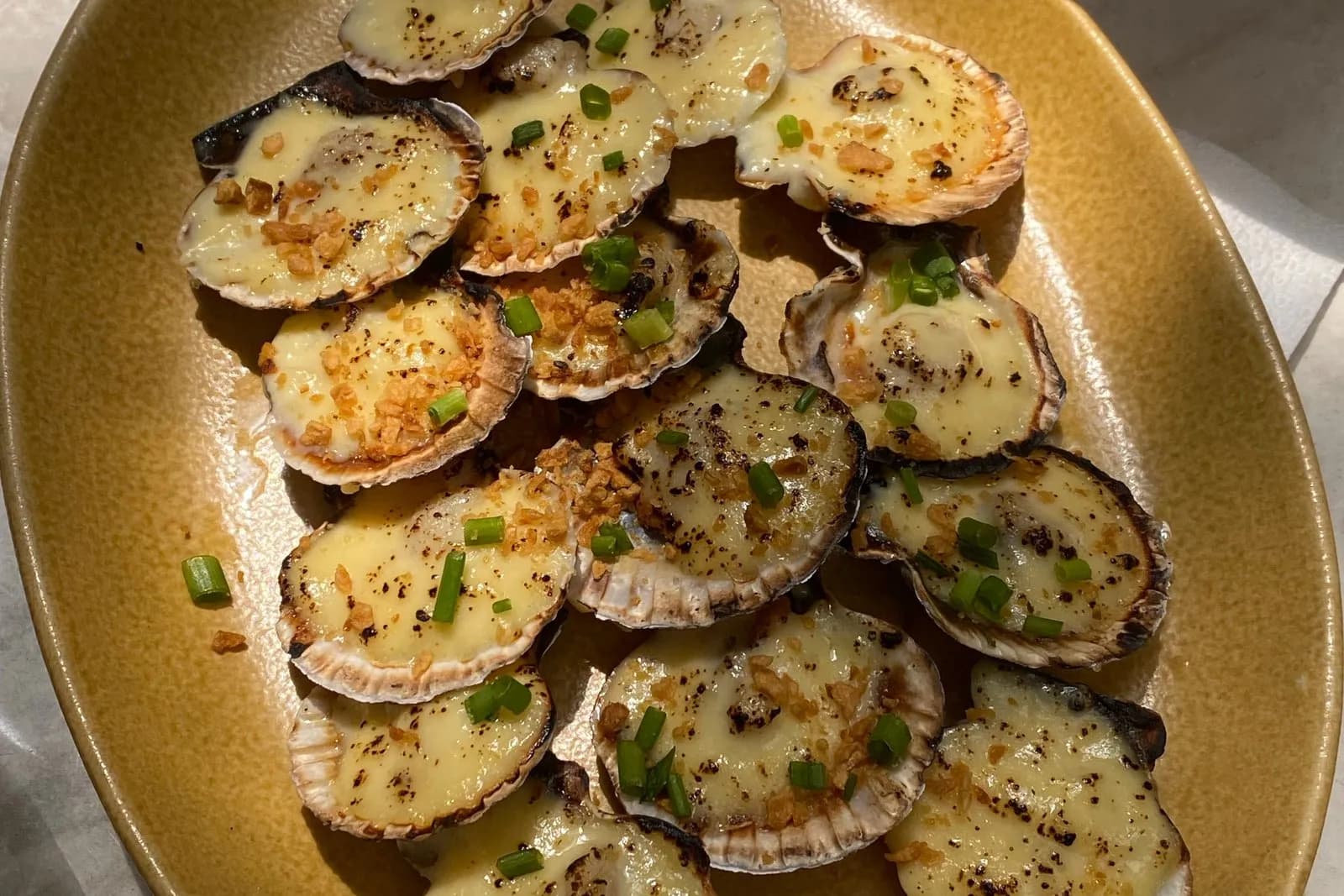 Vieiras gratinadas con queso fundido en sus conchas, más de una docena vistas desde arriba y solapadas sobre una fuente ovalada de cerámica ocre: cada una lleva una capa de queso de color crema, dorada y con motas chamuscadas, con ajo frito crujiente y cebolleta tierna picada por encima