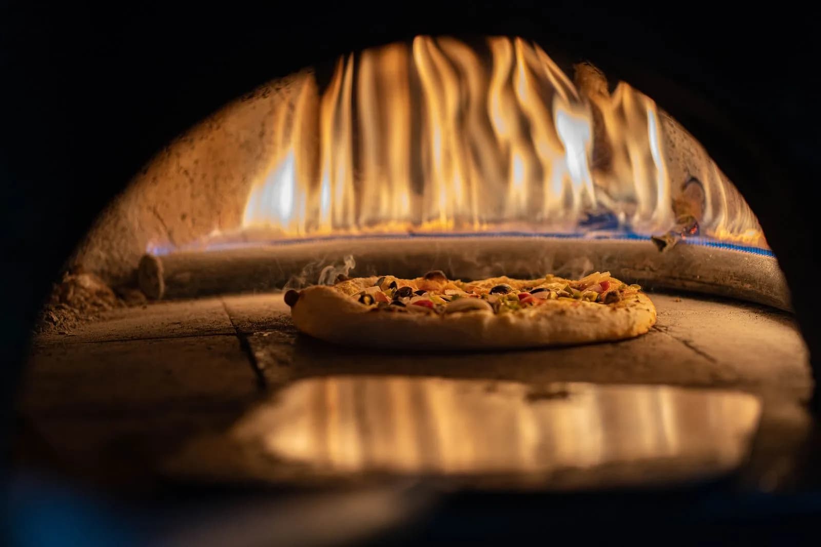 Pizza horneándose sobre la piedra de un horno de cúpula con llamas vivas al fondo y una pala de madera