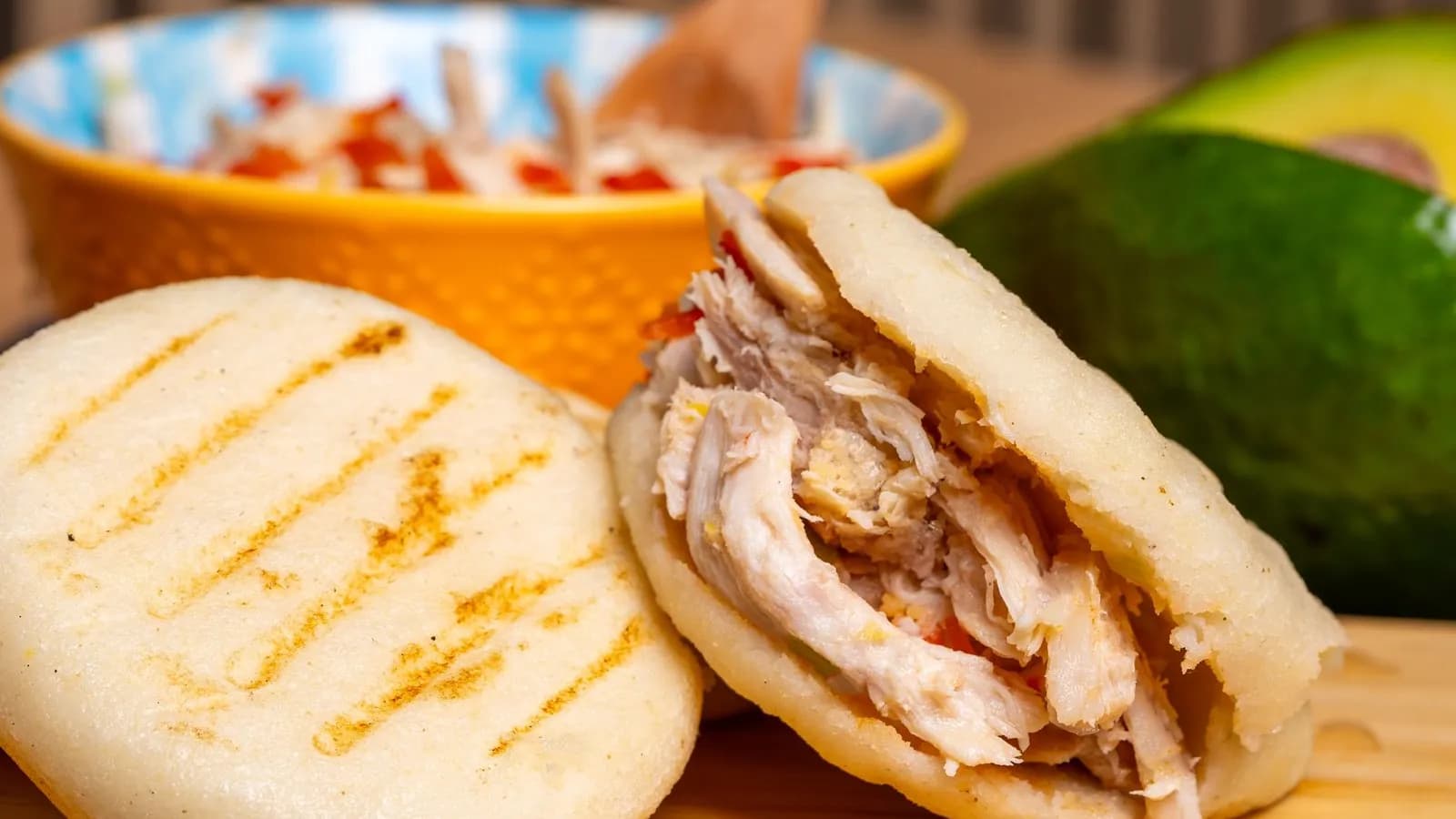 Dos arepas con rayas tostadas en la superficie: una entera y otra abierta por la mitad y rellena de pollo desmechado con trocitos de pimiento rojo; detrás, un cuenco naranja con más relleno y medio aguacate, sobre una tabla de madera clara