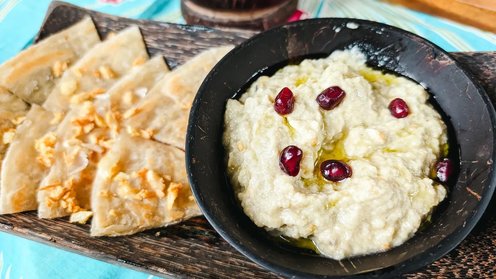 Cuenco de baba ganoush cremoso con granos de granada y aceite, junto a triángulos de pan de pita
