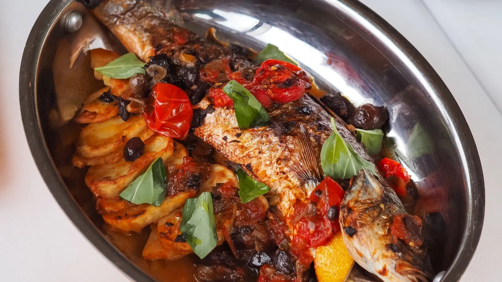Pescado entero asado, de piel tostada, sobre una cama de rodajas de patata asadas en una fuente ovalada de cobre con asas doradas, con tomates cherry, aceitunas negras, hojas de albahaca y un gajo de limón