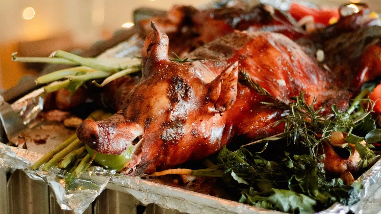 Cochinillo entero asado y servido en bandeja (no sale así del kamado), con la piel brillante y tostada y una manzana verde en la boca, sobre hojas verdes y romero en una bandeja de aluminio